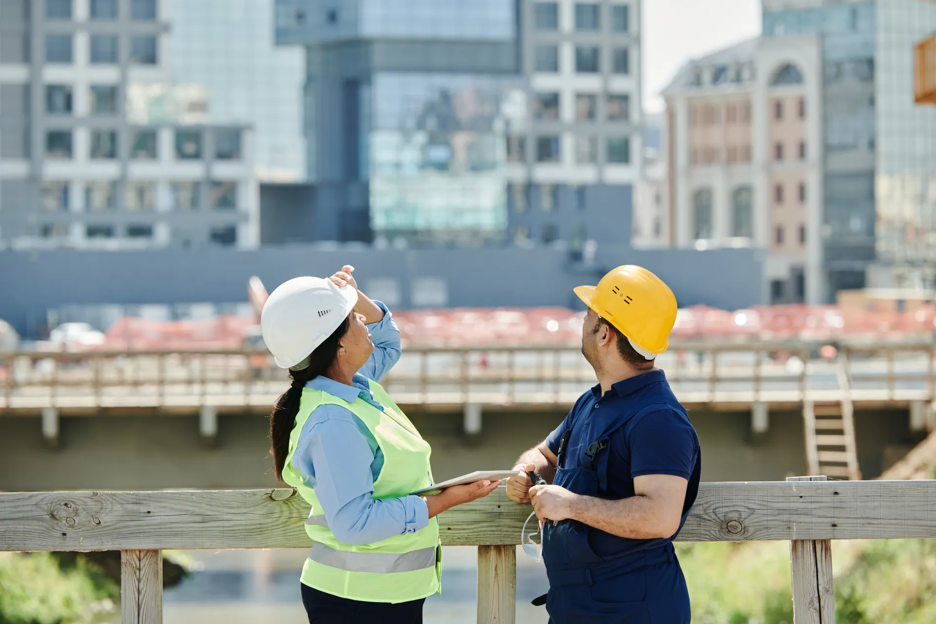 Two engineers in hard hats reviewing plans on a rooftop with city buildings behind
